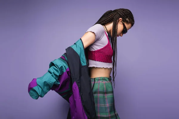 Vista laterale della donna con dreadlocks posa in occhiali da sole e giacca vintage isolato su viola — Foto stock