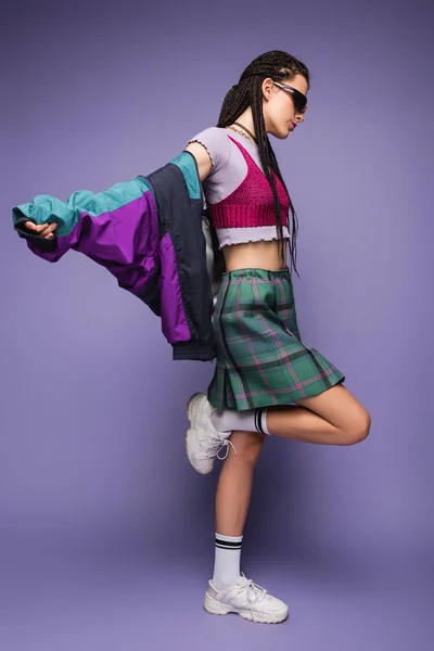 Side view of woman in sunglasses posing in nineties style clothes and white sneakers on purple background — Stock Photo