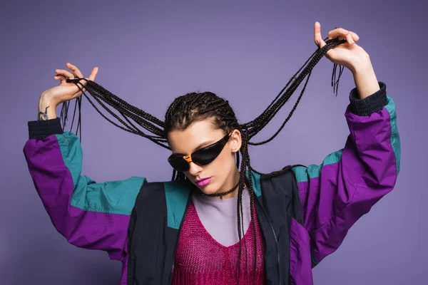 Stylish woman in sunglasses and vintage style jacket holding braided dreadlocks isolated on purple — Stock Photo