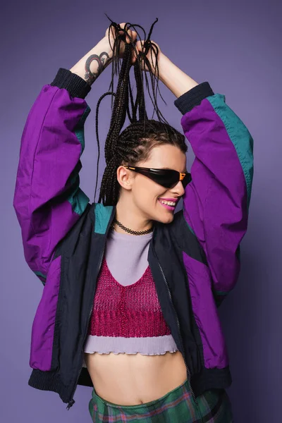 Mujer sonriente en gafas de sol y chaqueta vintage sosteniendo trenzas aisladas en púrpura - foto de stock
