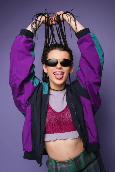 Giovane donna in giacca vintage e occhiali da sole che tengono le trecce e sporgono la lingua isolata sul viola — Foto stock