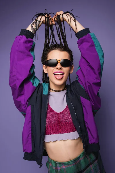 Mujer joven en chaqueta vintage y gafas de sol sosteniendo trenzas y sobresaliendo lengua aislada en púrpura - foto de stock