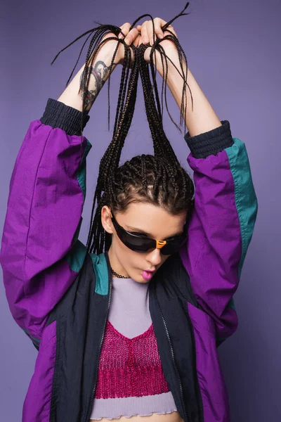 Брюнетка женщина в винтажном пиджаке и солнцезащитных очках держа косички и надувая губы изолированы на фиолетовый — стоковое фото