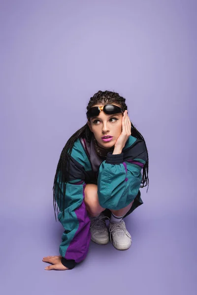 Femme avec dreadlocks tressés et lunettes de soleil assis sur des hanches dans les années nonante veste de style sur fond violet — Photo de stock