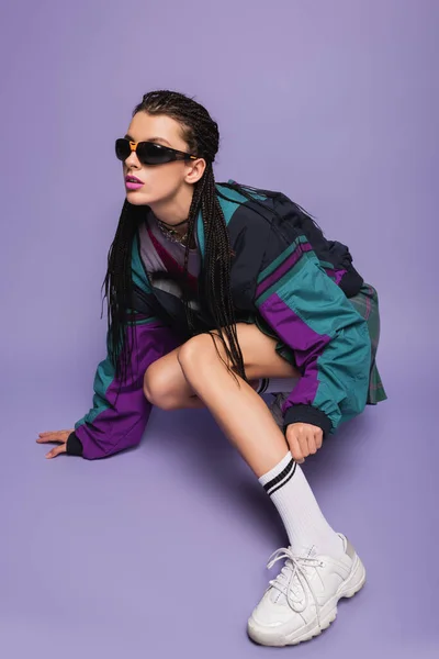 Young woman in sunglasses and vintage jacket adjusting white sock on purple background — Stock Photo