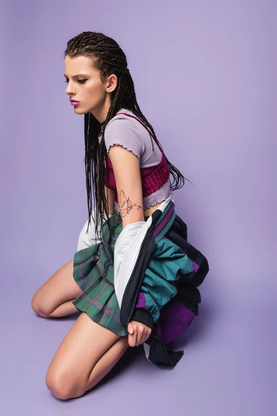 Стильная татуированная женщина с плетеными дредами на коленях на фиолетовом фоне — стоковое фото