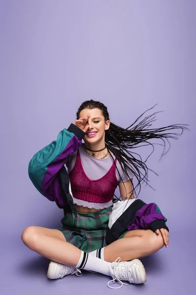 Smiling woman in nineties attire sitting with crossed legs on wind on purple background — Stock Photo