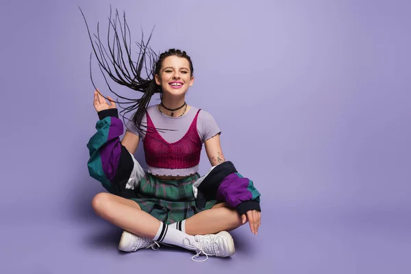 Smiling woman in vintage outfit waving hand while sitting on wind with crossed legs on purple background — Stock Photo