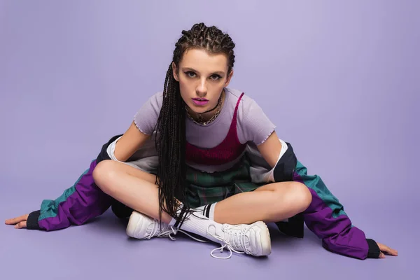 Woman with braids sitting in casual nineties attire with crossed legs and looking at camera on purple background — Stock Photo