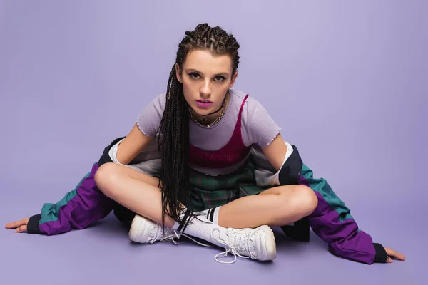 Mujer con trenzas sentado en traje casual de los noventa con las piernas cruzadas y mirando a la cámara en el fondo púrpura - foto de stock