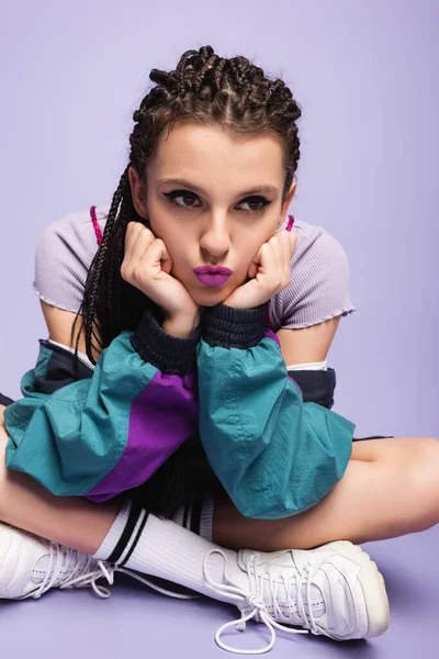 Thoughtful nineties style woman sitting with crossed legs and pouting lips on purple background — Stock Photo