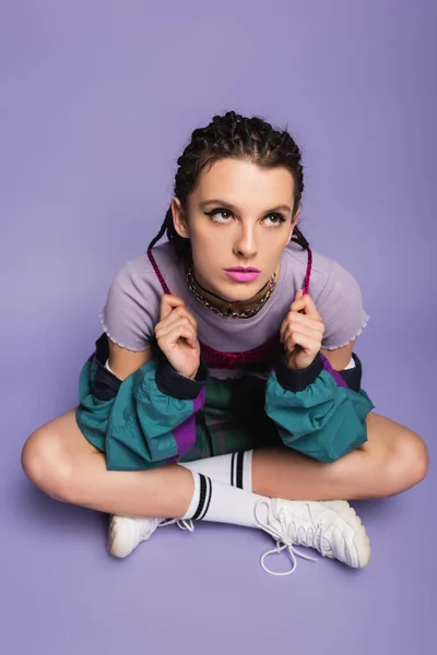 Mujer de estilo vintage sosteniendo trenzas mientras está sentado con las piernas cruzadas y mirando hacia otro lado sobre fondo púrpura - foto de stock