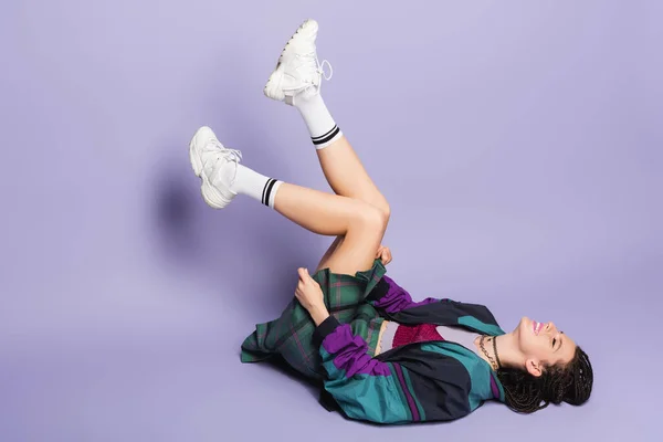 Excited nineties style woman in white sneakers lying with raised legs on purple background — Stock Photo