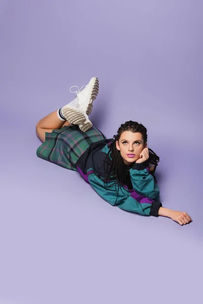 Dreamy woman in nineties style outfit and white sneakers lying and looking away on purple background — Stock Photo