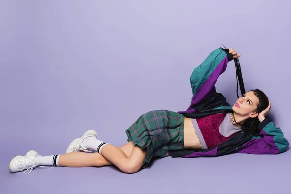 Задумчивая женщина в винтажной одежде и белых кроссовках, держащих плетеные дреды, лежа на фиолетовом фоне — стоковое фото