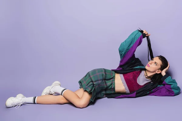 Pensive woman in vintage clothes and white sneakers holding braided dreadlocks while lying on purple background — Stock Photo