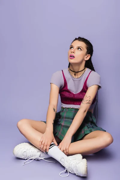 Amazed woman in plaid skirt and white sneakers sitting with crossed legs and looking away on purple background — Stock Photo