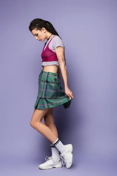 Side view of tattooed woman in plaid skirt and white sneakers standing on purple background — Stock Photo