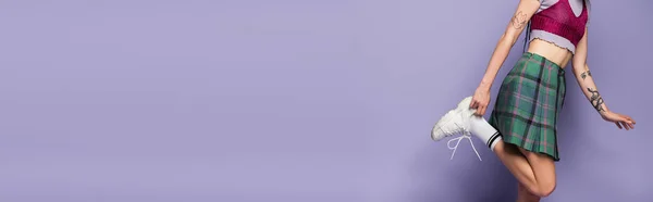 Обрезанный вид татуированной женщины в клетчатой юбке регулируя белые кроссовки на фиолетовом фоне, баннер — стоковое фото