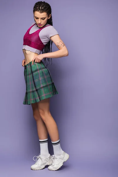 Full length of woman in crop top and white sneakers adjusting checkered skirt on purple background — Stock Photo