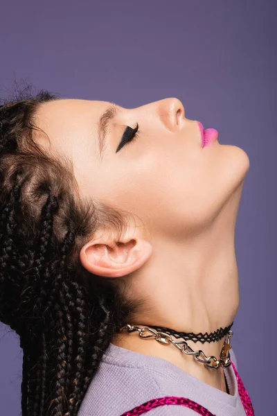 Perfil de mujer con maquillaje y rastas trenzadas posando con la cabeza levantada aislada en púrpura - foto de stock