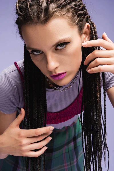 Giovane donna con trucco trecce toccanti e guardando la fotocamera isolata su viola — Foto stock