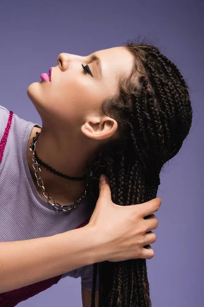 Retrato de mujer con estilo con maquillaje sosteniendo rastas trenzadas aisladas en púrpura - foto de stock