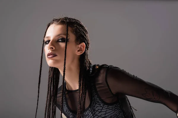 Mujer morena con estilo con maquillaje y trenzas mirando a la cámara aislada en gris - foto de stock