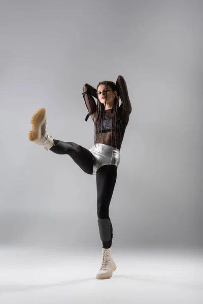 Full length of woman in black and tight jumpsuit and silver shorts posing on one leg on grey background — Stock Photo