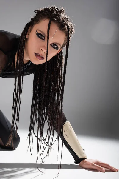 Futuristische Frau mit stylischem Make-up und geflochtenen Dreadlocks, die auf weißer Oberfläche und grauem Hintergrund in die Kamera schauen — Stockfoto