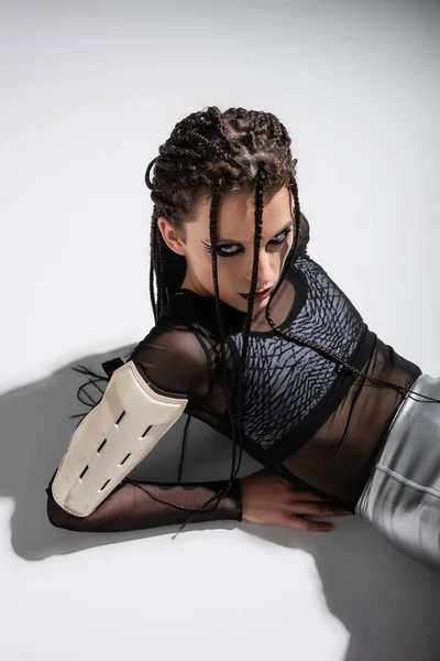 Brunette woman with braids and expressive makeup lying in futuristic outfit on grey background — Stock Photo