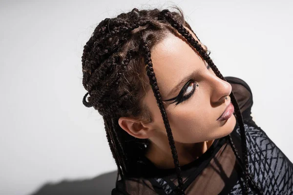 Portrait of young woman with braids and futuristic makeup looking away on grey background — Stock Photo