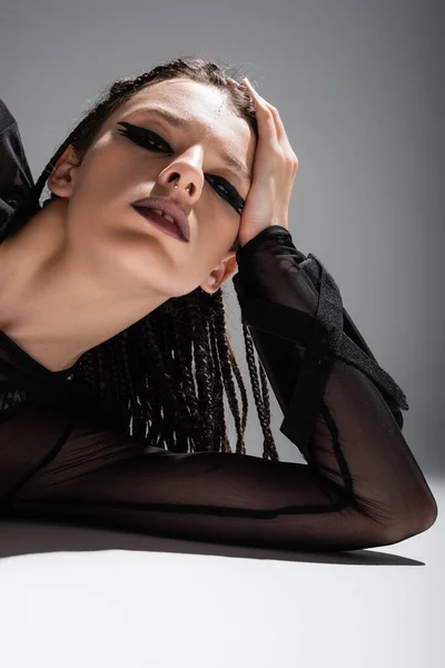 Femme avec dreadlocks tressés et maquillage futuriste avec piercing couché et regardant la caméra sur fond gris — Photo de stock