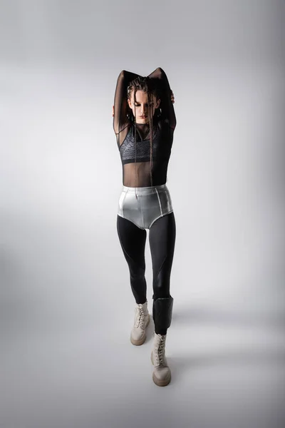 Full length of woman in silver shorts over black tight jumpsuit standing with hands behind head on grey background — Stock Photo