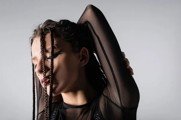 Mujer elegante con trenzas y ojos cerrados posando con las manos detrás de la cabeza aisladas en gris - foto de stock