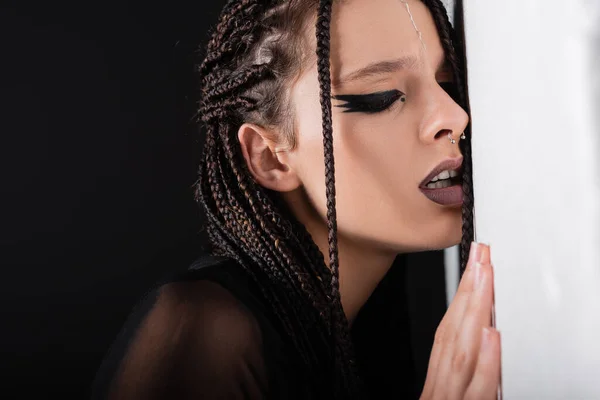 Sensual woman with braids and futuristic makeup near while wall on black background — Stock Photo