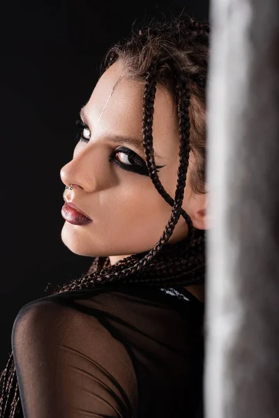 Retrato de mulher com dreadlocks e maquiagem elegante olhando para a câmera perto de parede borrada no fundo preto — Fotografia de Stock