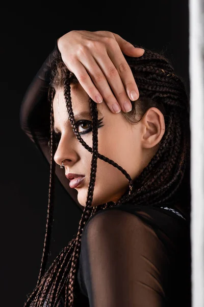 Porträt einer stilvollen Frau mit Make-up und piercing berühren Zöpfe in der Nähe der weißen Wand auf schwarzem Hintergrund — Stockfoto