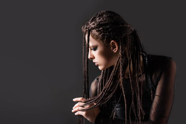 Giovane donna in abiti oscuri futuristici che toccano dreadlocks mentre posa isolata sul nero — Foto stock