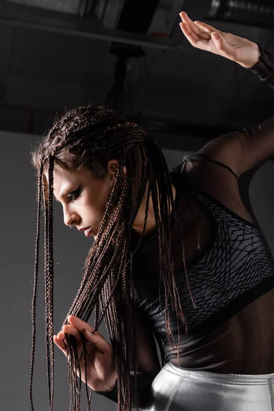 Mujer futurista en traje negro tocando rastas trenzadas mientras posa sobre fondo gris - foto de stock