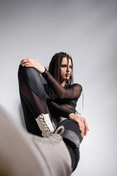 Trendy woman with dreadlocks sitting in black futuristic outfit on grey background — Stock Photo