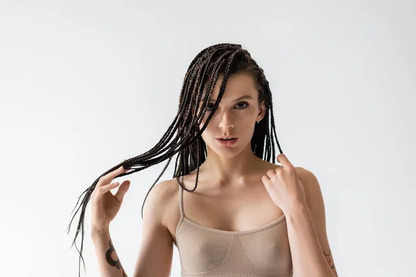 Young woman in beige top touching braids while looking at camera isolated on grey — Stock Photo