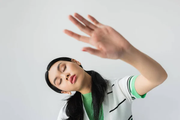 Pretty asian model closing eyes and outstretching blurred hand isolated on grey — Stock Photo