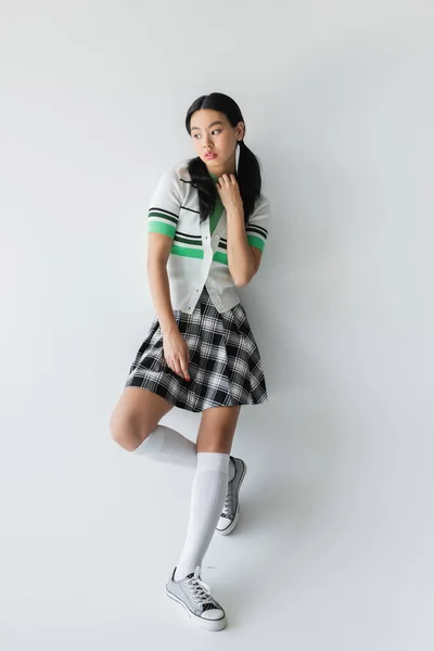 Mujer asiática con estilo en cárdigan recortado y zapatillas de deporte posando cerca de fondo gris - foto de stock