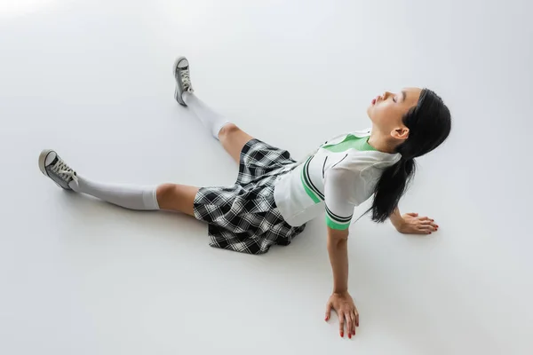 Side view of young asian model in plaid skirt sitting on grey background — Stock Photo