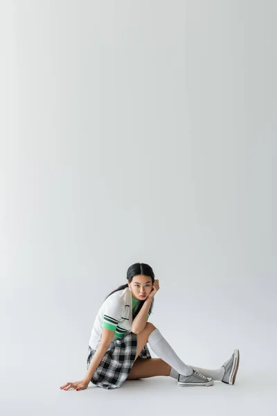 Trendy asian model in cropped cardigan looking at camera while sitting on grey background — Stock Photo