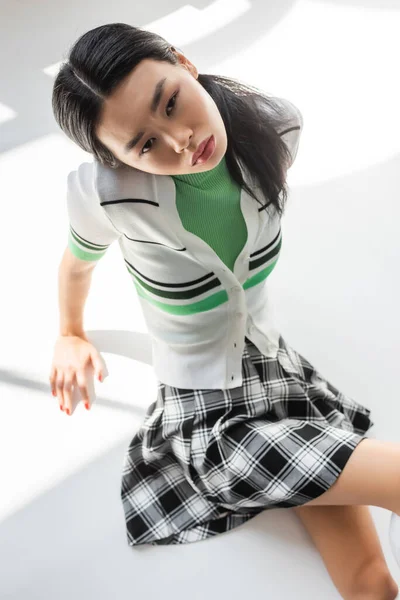 Hochwinkelaufnahme des trendigen asiatischen Modells, das auf grauem Hintergrund mit Licht in die Kamera blickt — Stockfoto