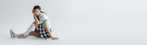 Fashionable asian woman in skirt and cropped cardigan sitting on grey background with copy space, banner — Stock Photo
