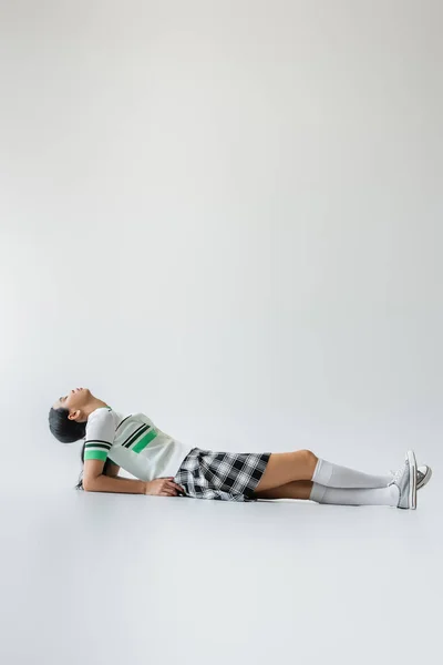 Vue latérale de la jeune femme asiatique en chaussettes de genou et jupe couchée sur fond gris — Photo de stock