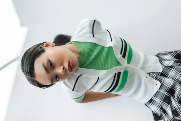 Draufsicht einer jungen Asiatin in abgeschnittener Strickjacke, die in die Kamera auf grauem Hintergrund blickt — Stockfoto